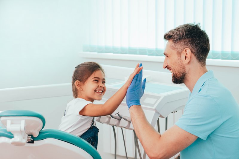 Little girl high fiving dentist's gloved hand as he squats to her height in exam room